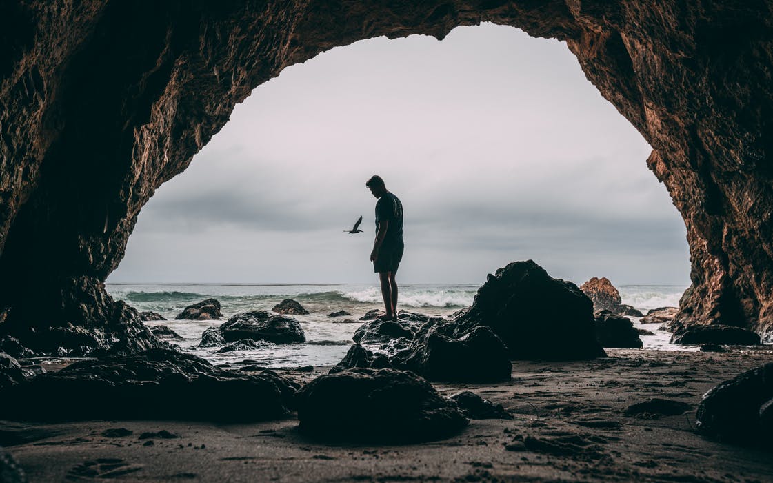 Photo of man standing on rock at entrance to cave near shore